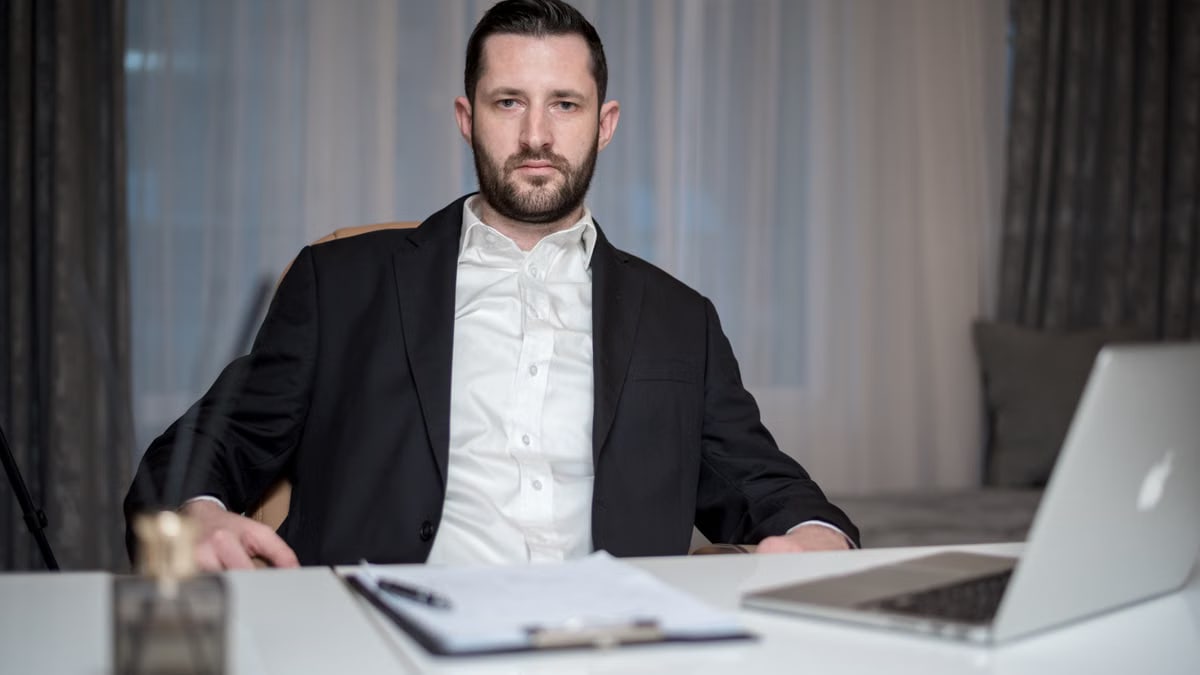 A serious bearded man in a black suit sits behind a desk with a laptop and documents.